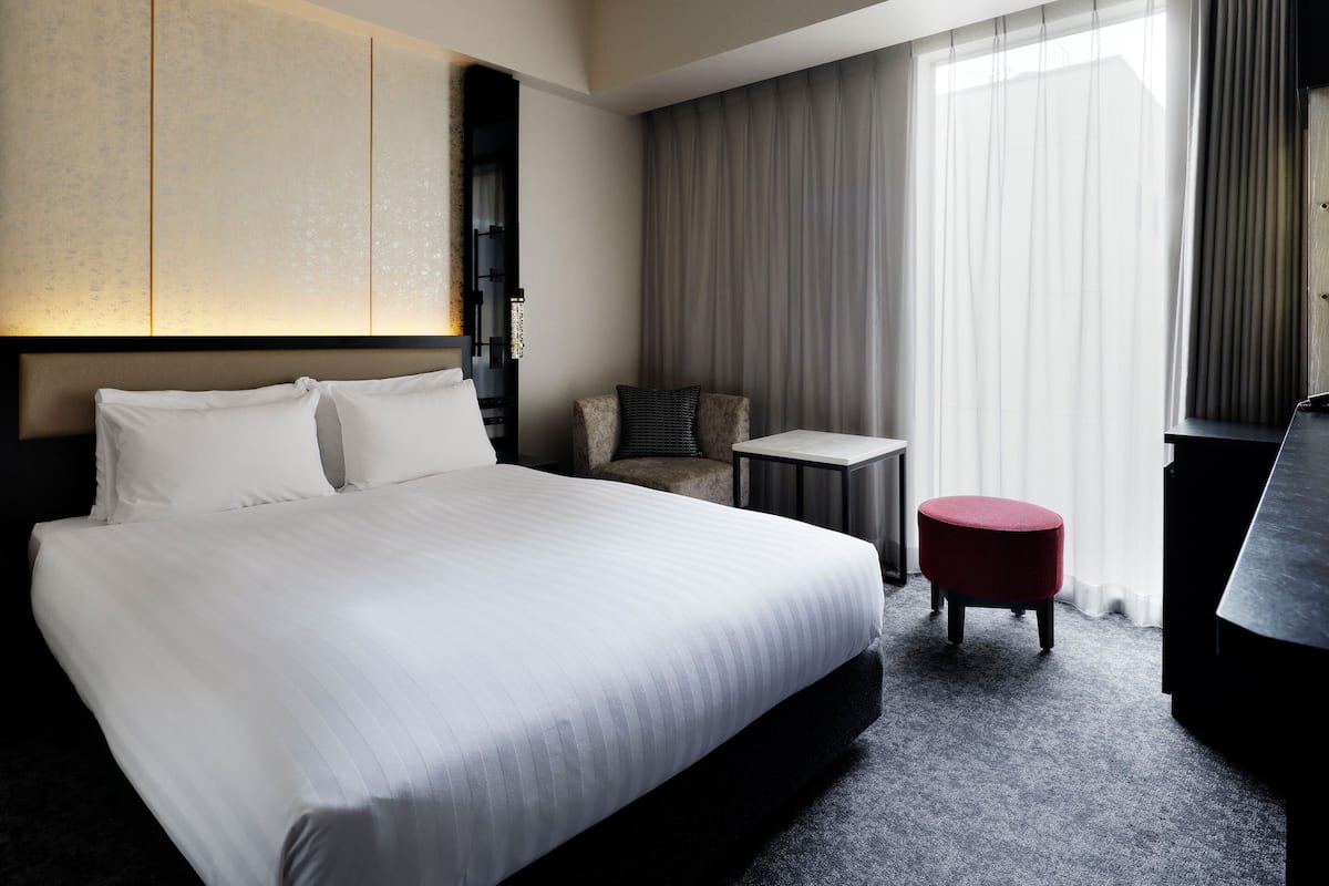 Une chambre d’hôtel avec un grand lit, une petite table, un pouf rouge, un canapé et une grande fenêtre avec des rideaux.