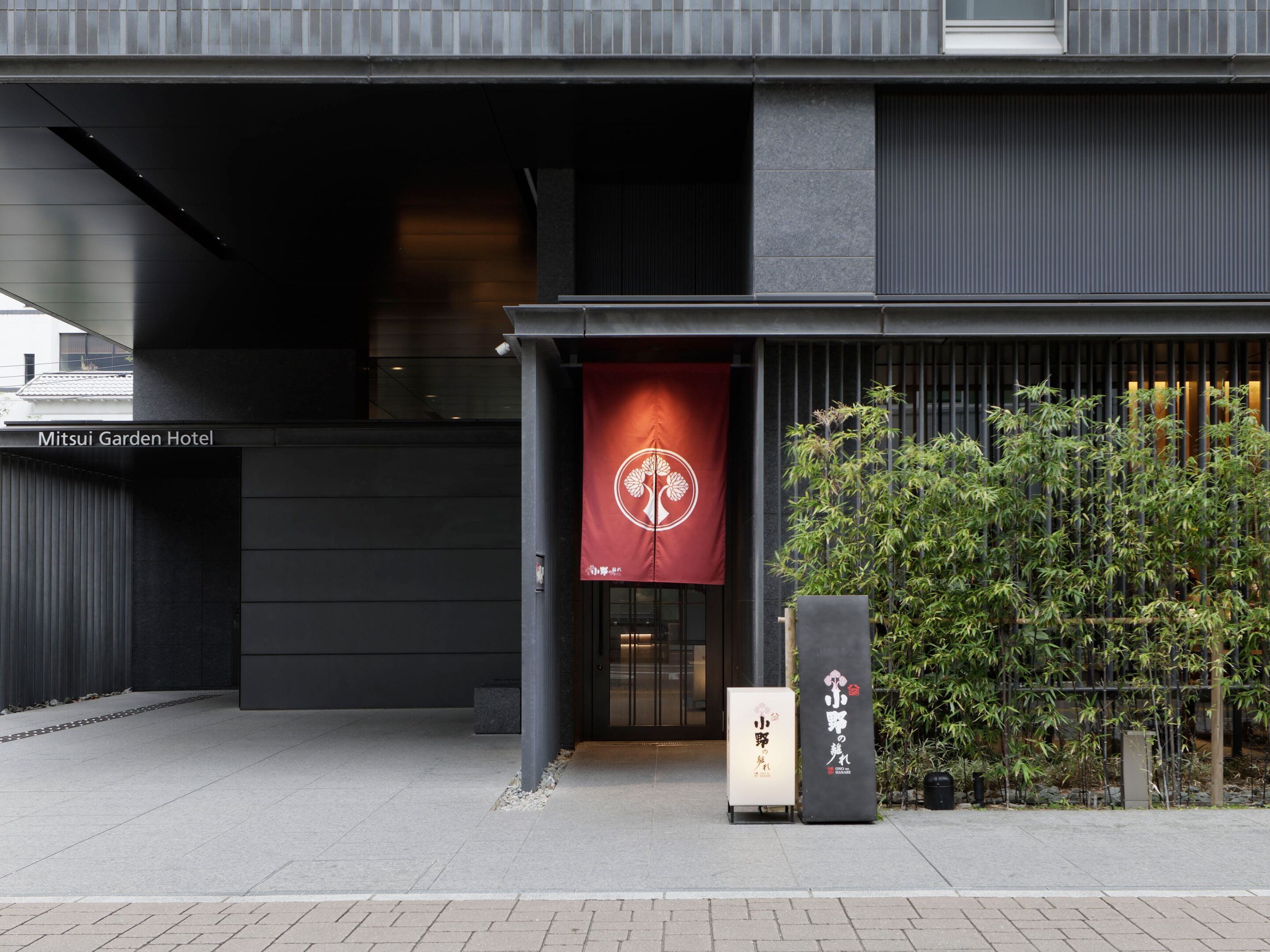 Entrada de um prédio com uma faixa vermelha, uma placa indicando 'Mitsui Garden Hotel' e uma pequena placa com caracteres japoneses.