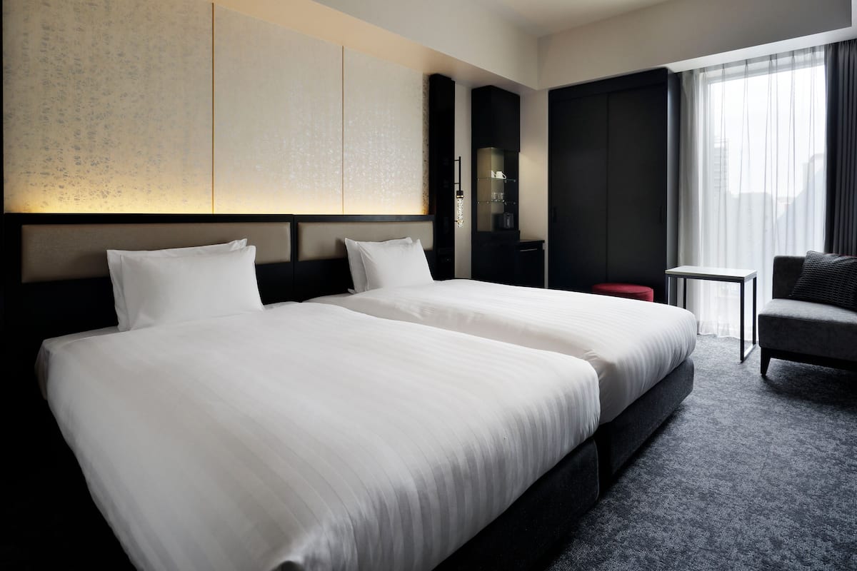 Une chambre d’hôtel avec deux lits, une petite table, une chaise et une grande fenêtre avec des rideaux.