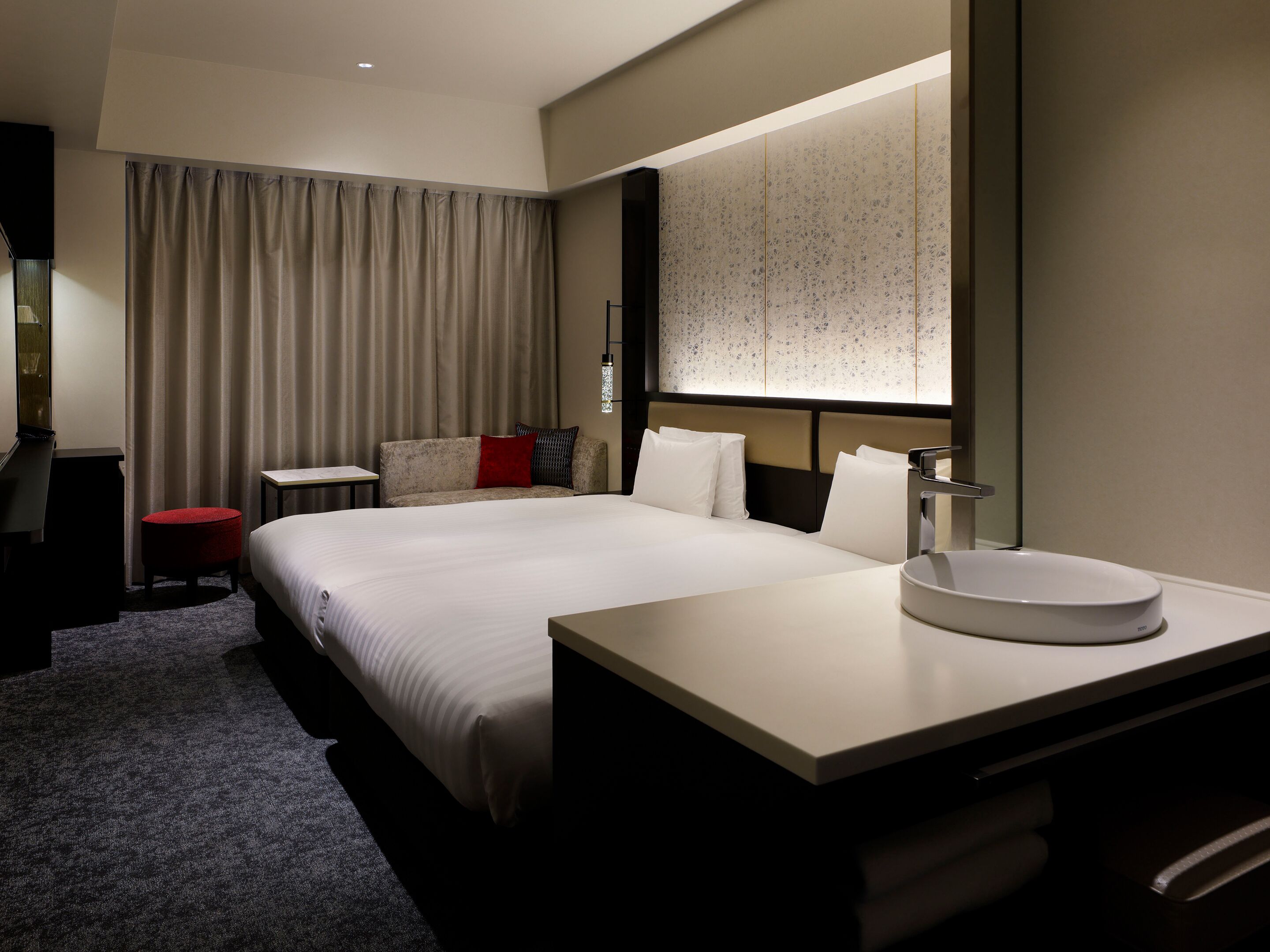 A hotel room with a large bed, a bedside table, a round mirror, and a small red stool.