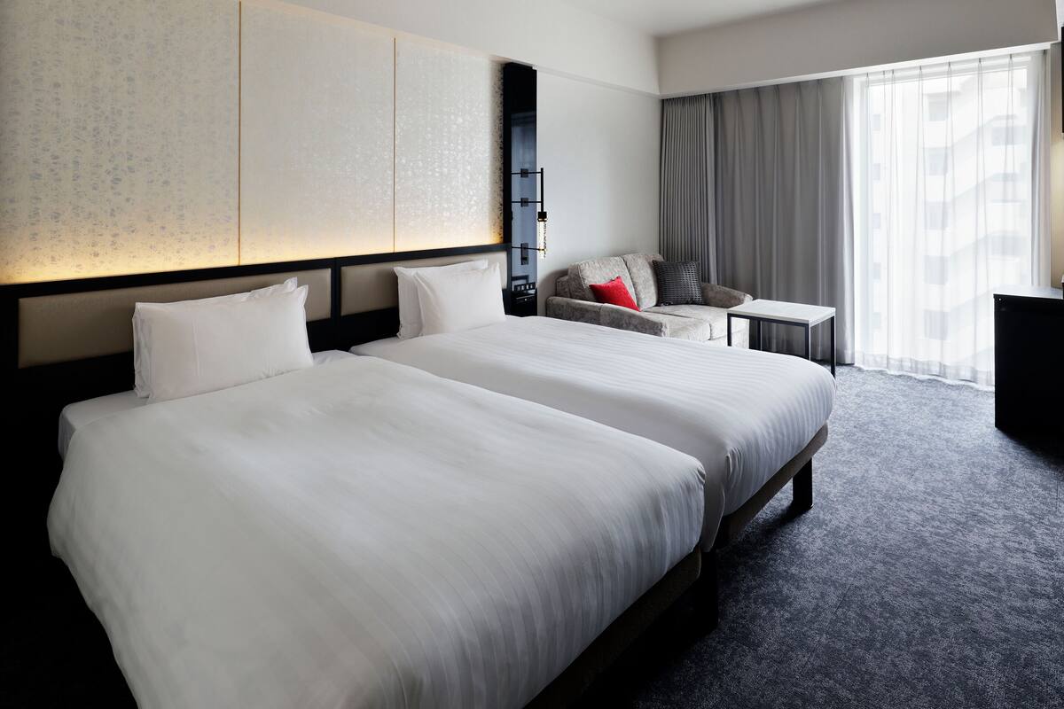 Une chambre d’hôtel avec deux grands lits, un petit bureau, un canapé et une grande fenêtre avec des rideaux.