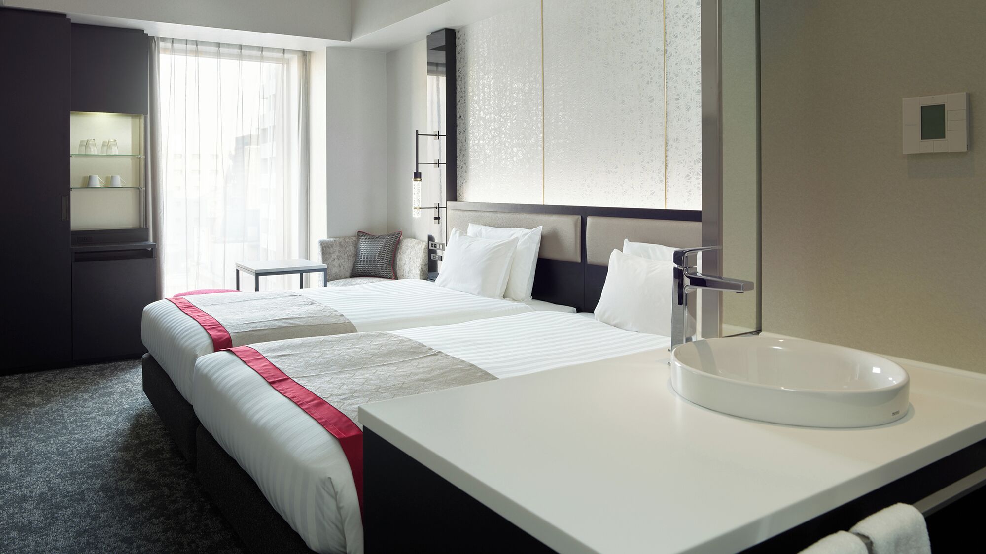 A hotel room with two beds, a large mirror, a round sink, and a window with sheer curtains.