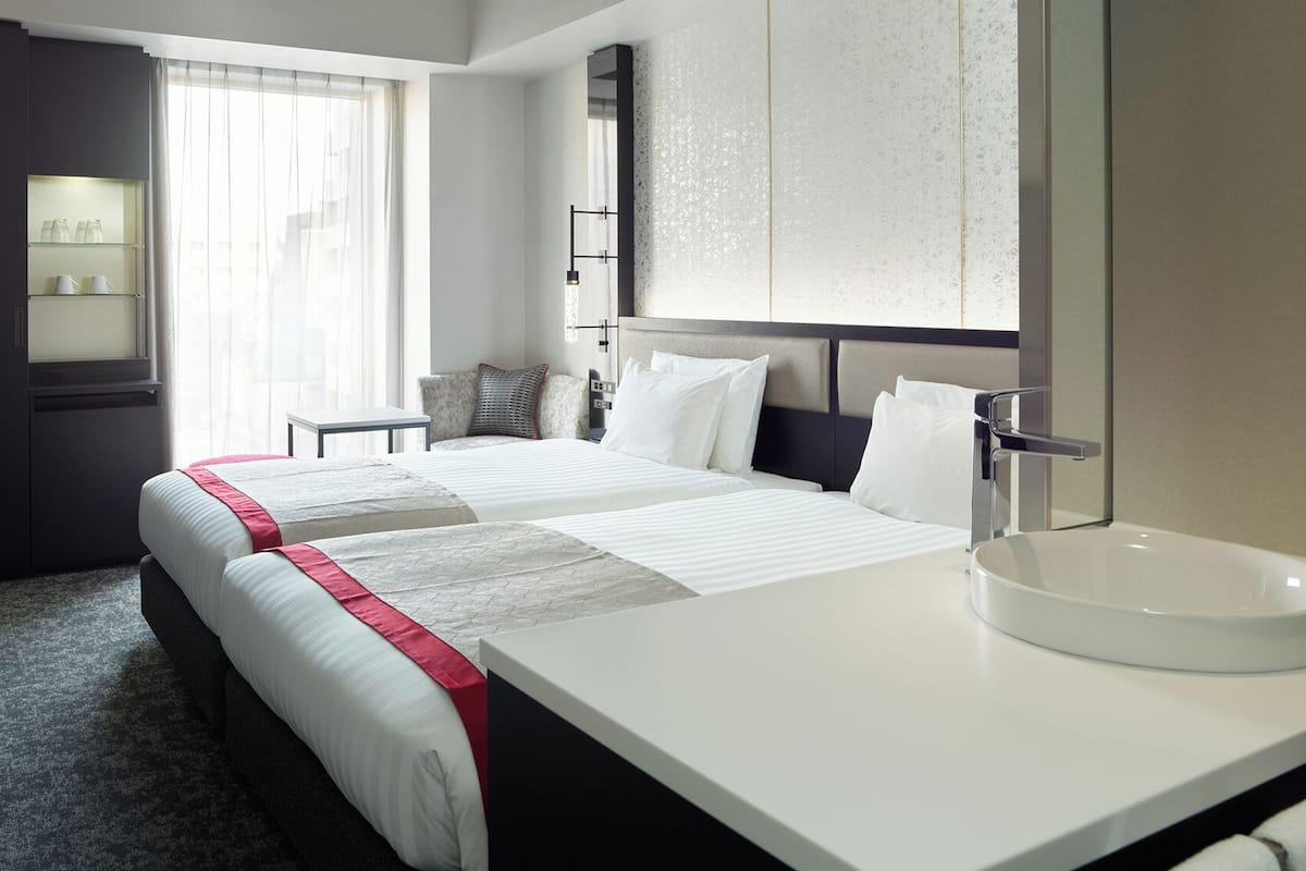 A hotel room with two beds, a large mirror, a round sink, and a window with sheer curtains.
