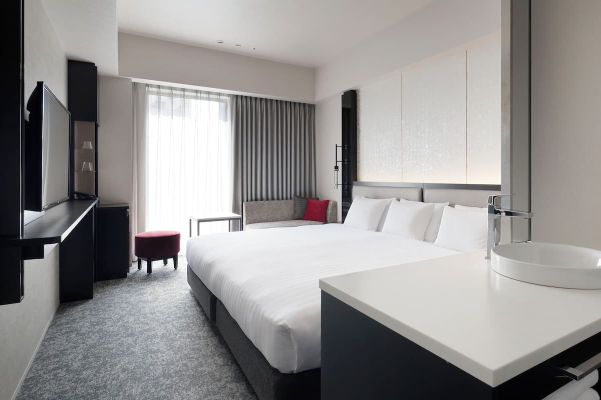 Une chambre d’hôtel avec un grand lit, un bureau, une chaise, un petit pouf, une salle de bain avec un lavabo et un miroir.