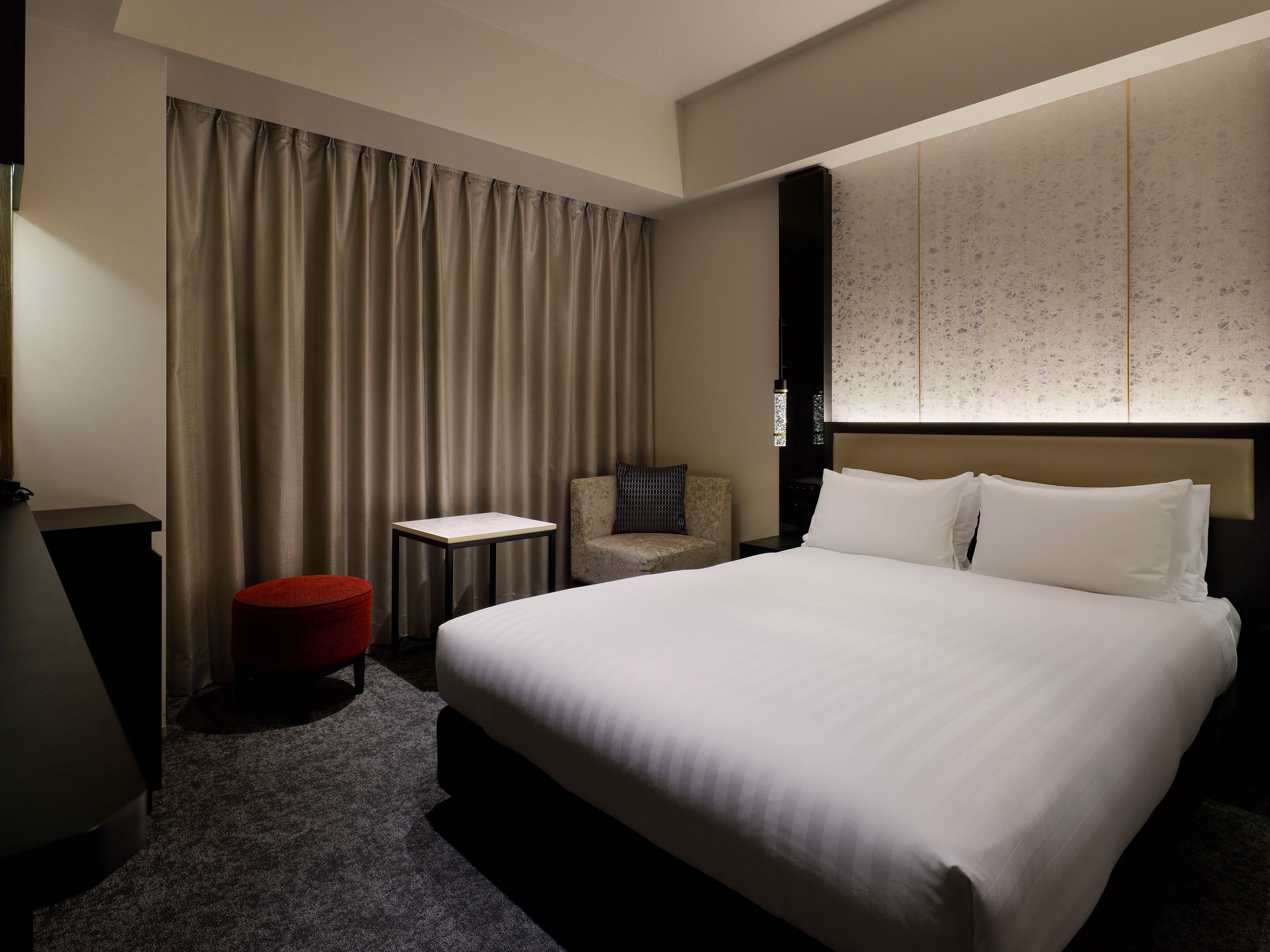 Habitación de hotel con una cama grande, una mesita pequeña, una silla y un reposapiés rojo.