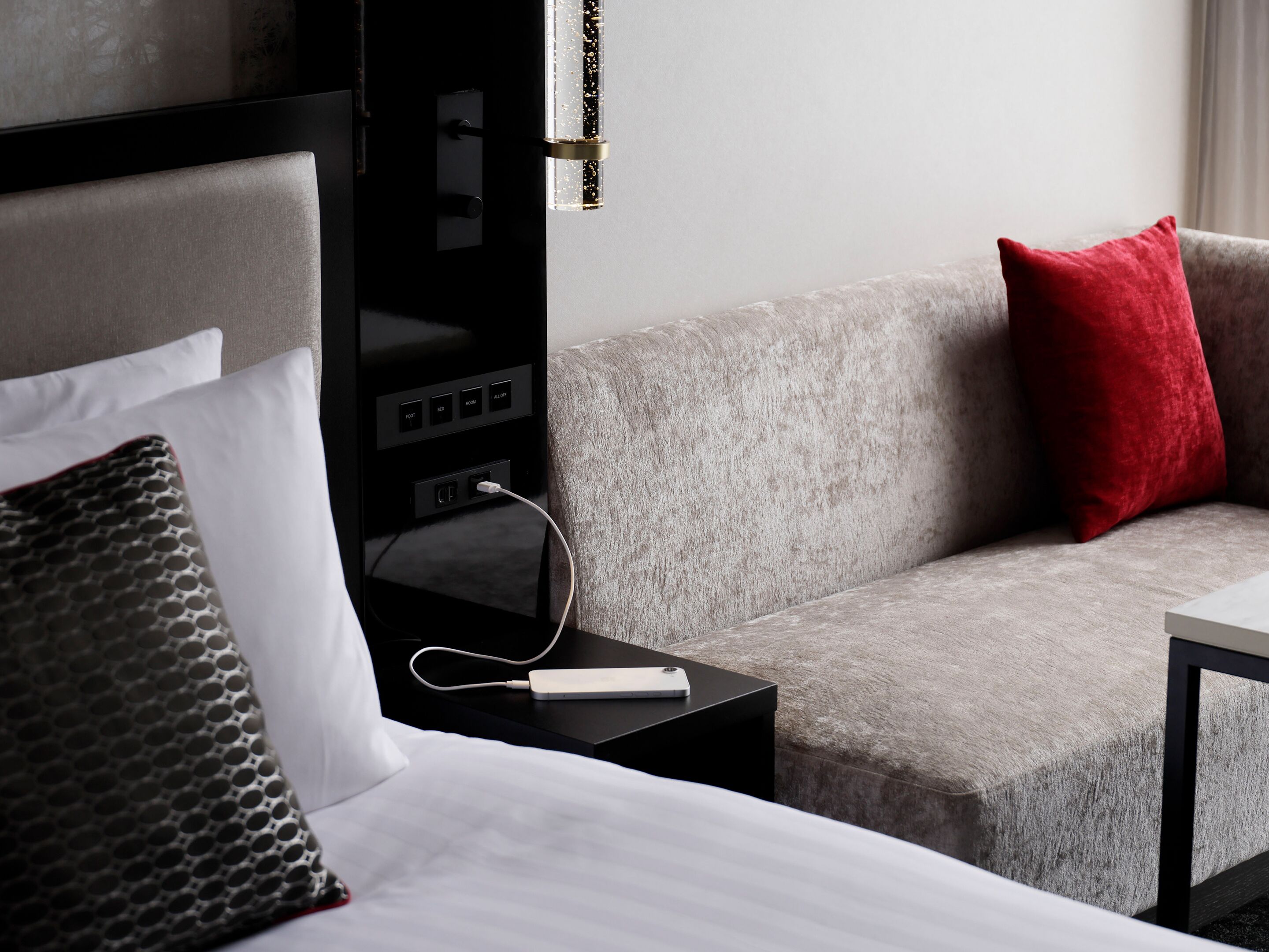 Una habitación de hotel con una cama, un sofá gris con un cojín rojo, una mesita de noche negra con un dispositivo blanco y un mueble negro para televisión con un enchufe.