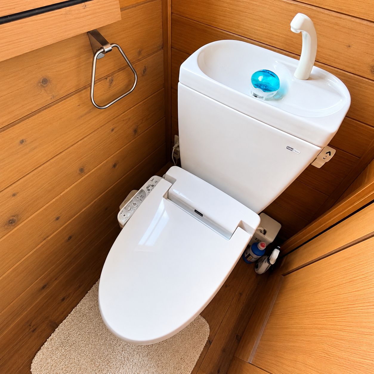 A compact, wooden-paneled bathroom with a white toilet, a blue water bottle, and a metal toilet paper holder.
