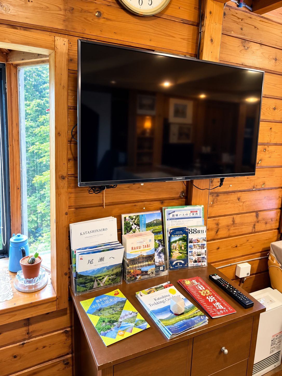 Interior de madera con un televisor de pantalla plana montado en la pared, una ventana con vistas a zonas verdes y una mesa con folletos turísticos y un control remoto.