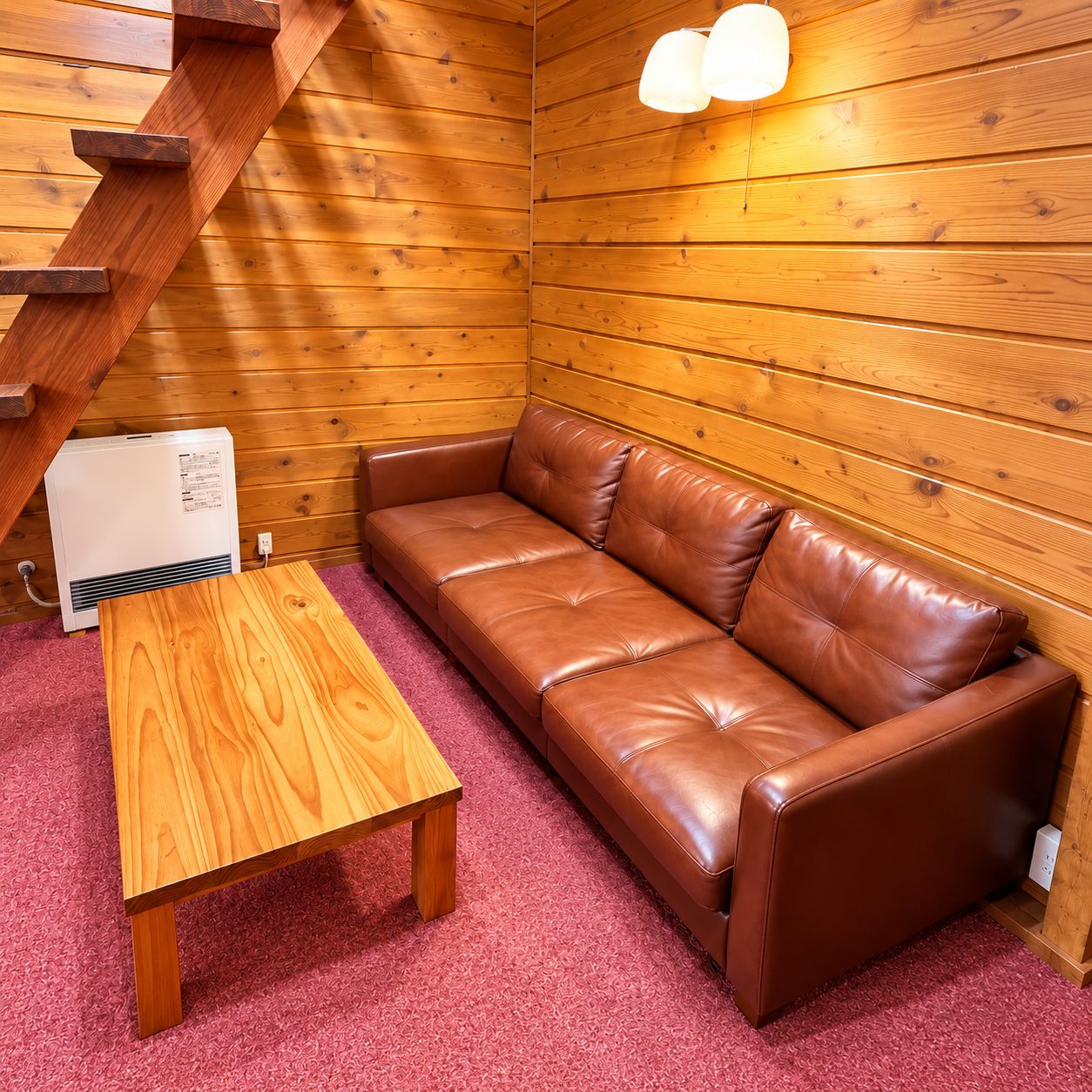 Interior de una cabaña de madera con un sofá de cuero marrón, una mesa de madera y una escalera.