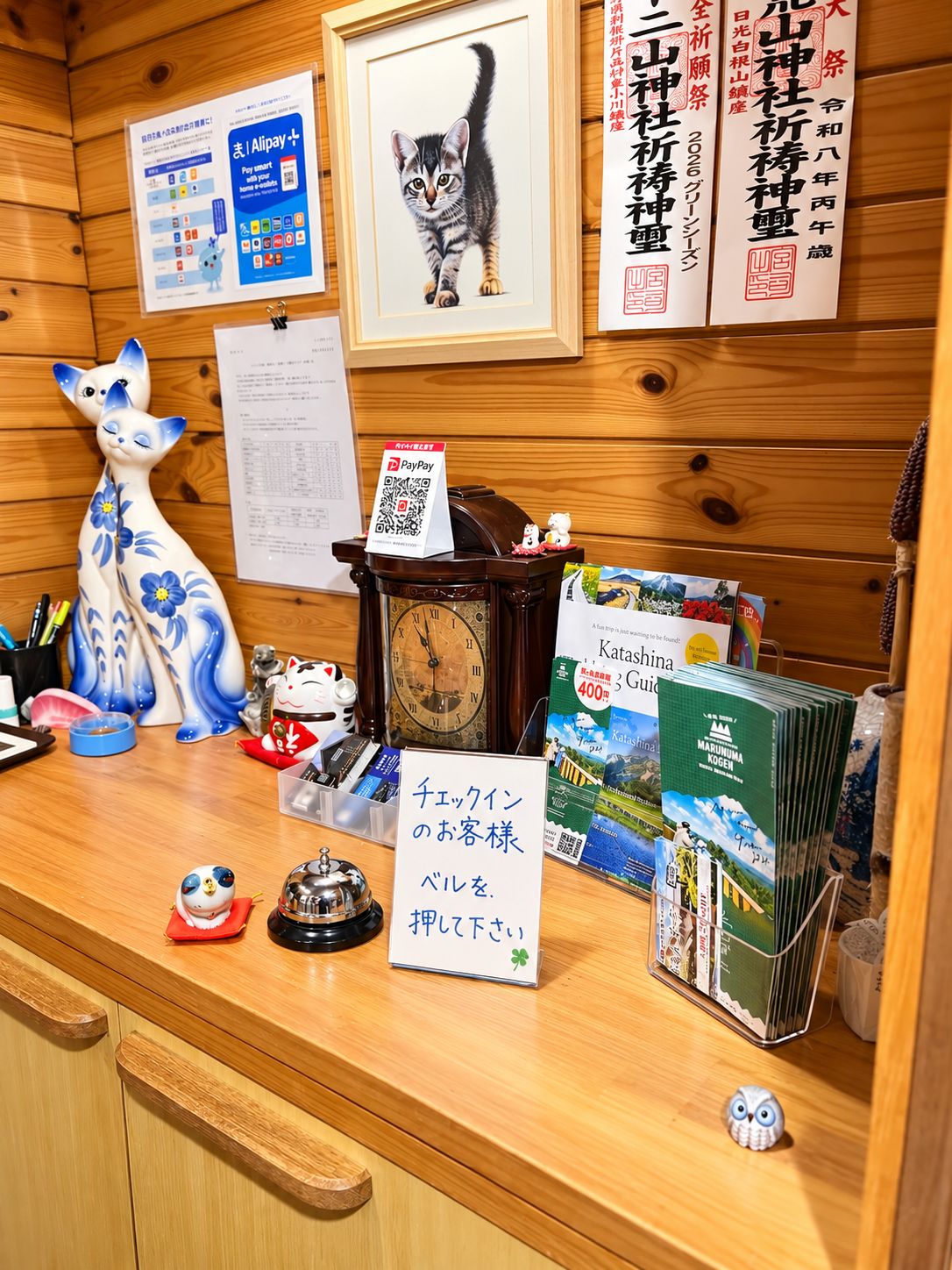 Um balcão de recepção de madeira com um relógio, uma fotografia de um gato emoldurada, uma placa com código QR e folhetos de viagens.
