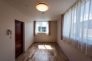 Un pasillo con suelo de madera, una ventana con cortinas translúcidas, una puerta y una lámpara en el techo.