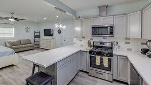 Uma cozinha moderna com armários brancos, um fogão de aço inoxidável e um micro-ondas. Há uma área de refeições com uma mesa branca e um sofá.