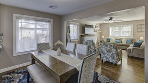 Uma área de refeições com uma mesa de madeira, cadeiras estampadas e um vaso de flores. Há uma sala de estar com um sofá e um ventilador de teto.