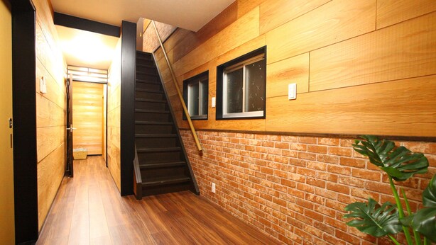 A narrow hallway with wooden flooring and walls, leading to a staircase with a wooden handrail and a black railing. - Samy's Guest House (Osaka)