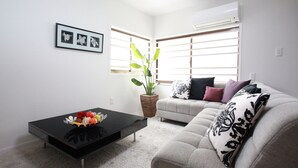 A living room with a black coffee table, a grey sofa, a potted plant, and a window with blinds. - Samy's Guest House (Osaka)