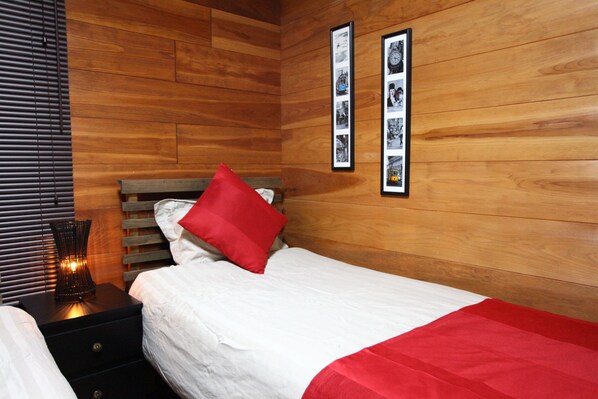 A bedroom with a wooden wall, a bed with white bedding and a red pillow, a nightstand with a lamp, and two framed pictures on the wall.