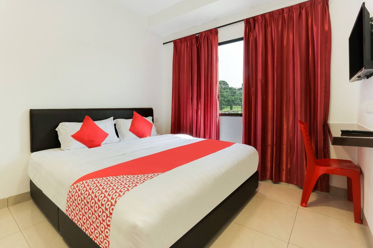 A hotel room with a bed, red curtains, a red chair, and a TV mounted on the wall.