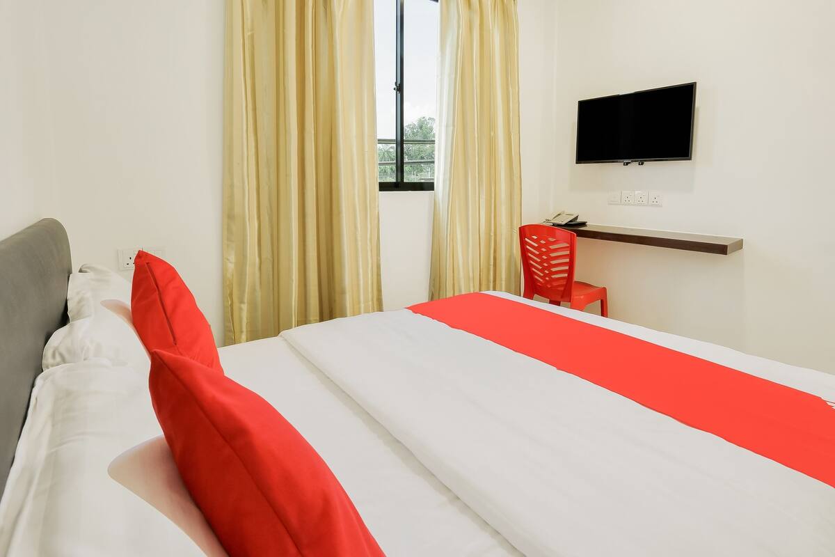 A hotel room with a bed, a TV mounted on the wall, a window with curtains, and a small desk with a chair.