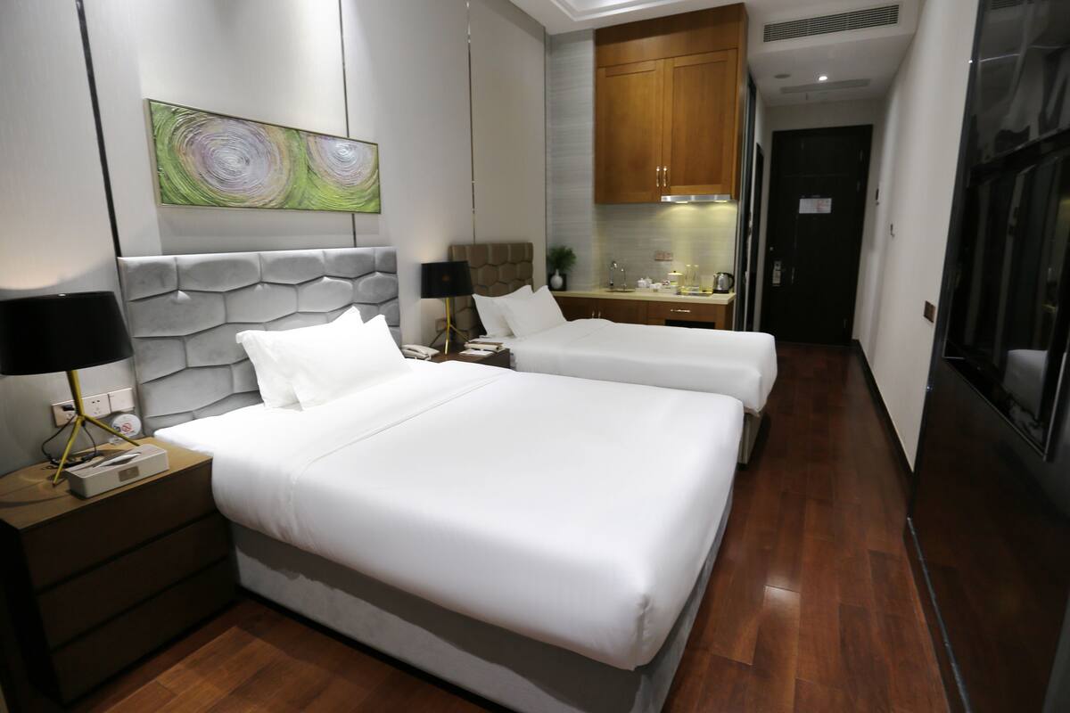 A hotel room with two beds, a nightstand with a lamp, a painting on the wall, and a kitchenette in the background.