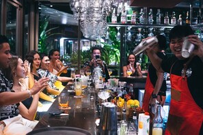 A bar area with a bartender preparing drinks, a long counter with various bottles and glasses, and a group of people seated at the bar. - Kata Gardens 4B Seaview Near Kata Noi (Karon)