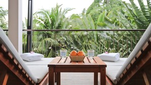A balcony with two lounge chairs, a small table with a bowl of fruit and glasses, and a view of lush greenery.