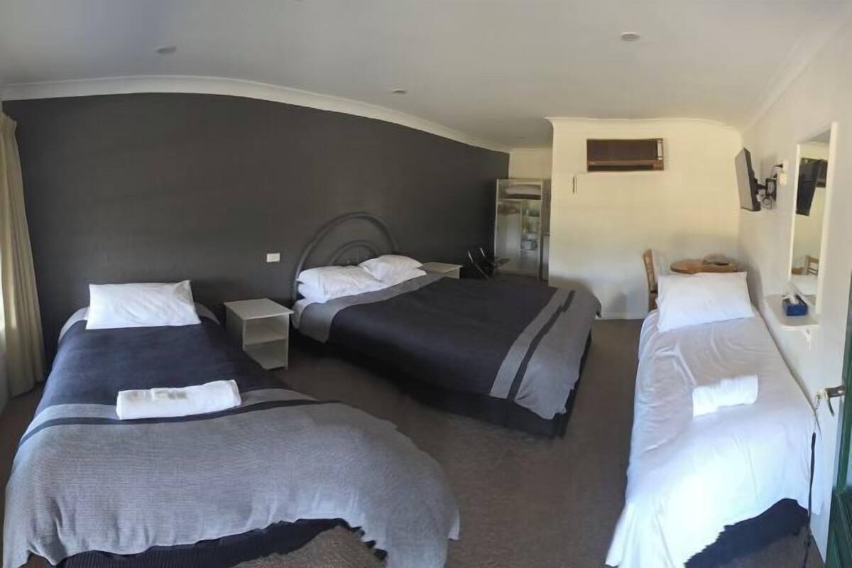 Quarto de hotel com três camas, uma escrivaninha pequena, um espelho e um guarda-roupa.