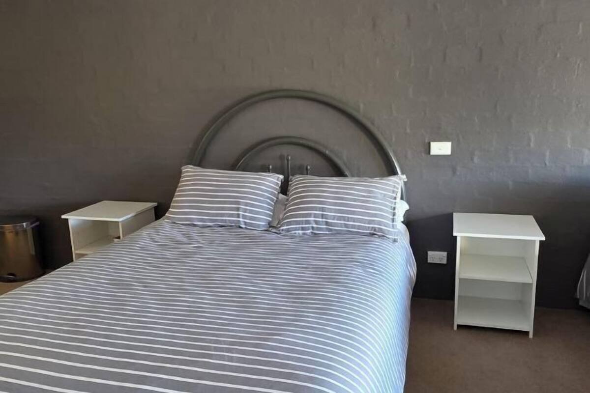 Uma cama com roupa de cama listrada, duas mesas de cabeceira, um criado-mudo branco e uma parede de tijolos cinzentos.