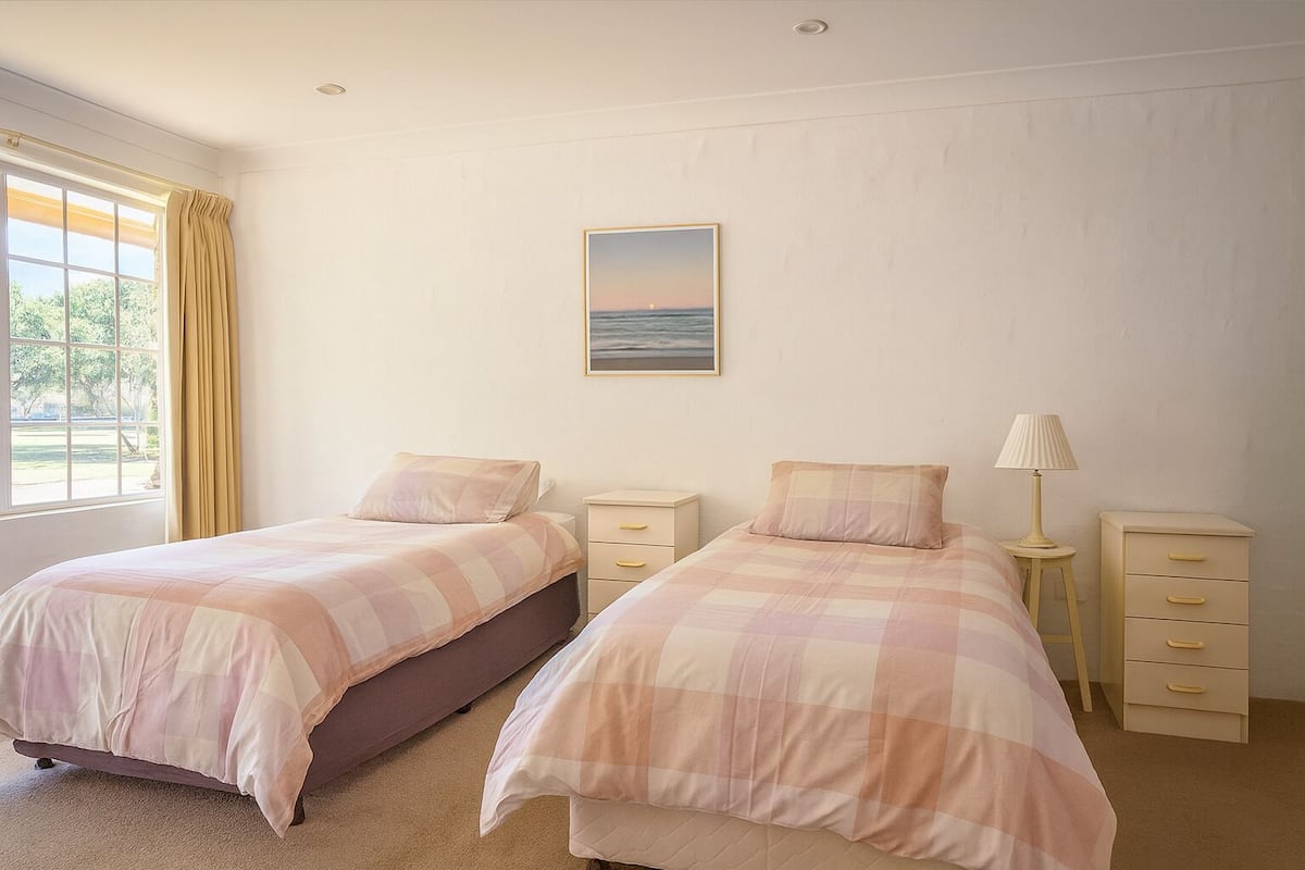 Quarto com duas camas, uma mesa de cabeceira com abajur, cômoda e janela com cortinas.