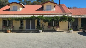 Un bâtiment à deux étages avec un toit rouge, un patio couvert agrémenté de chaises et de plantes en pot, et une allée gravillonnée.