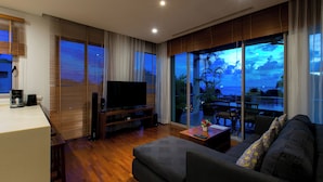 A modern living room with a flat-screen TV, wooden flooring, and a balcony with a view of the sky and palm trees. - Kata Gardens Rooftop Plunge Pool Penthouse 4C (Karon)