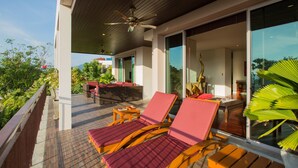 Un balcon doté de chaises longues en bois et d’une table, offrant une vue sur un jardin luxuriant et un bâtiment moderne.