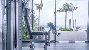Ein Fitnessraum mit Langhantelbank, Kurzhanteln und Klimmzugstange, mit Blick auf eine Holzterrasse, Palmen und den Ozean.