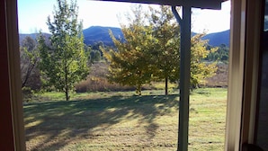 Vista desde una terraza cubierta con piso de madera, con vistas a un campo verde con árboles y montañas a lo lejos.