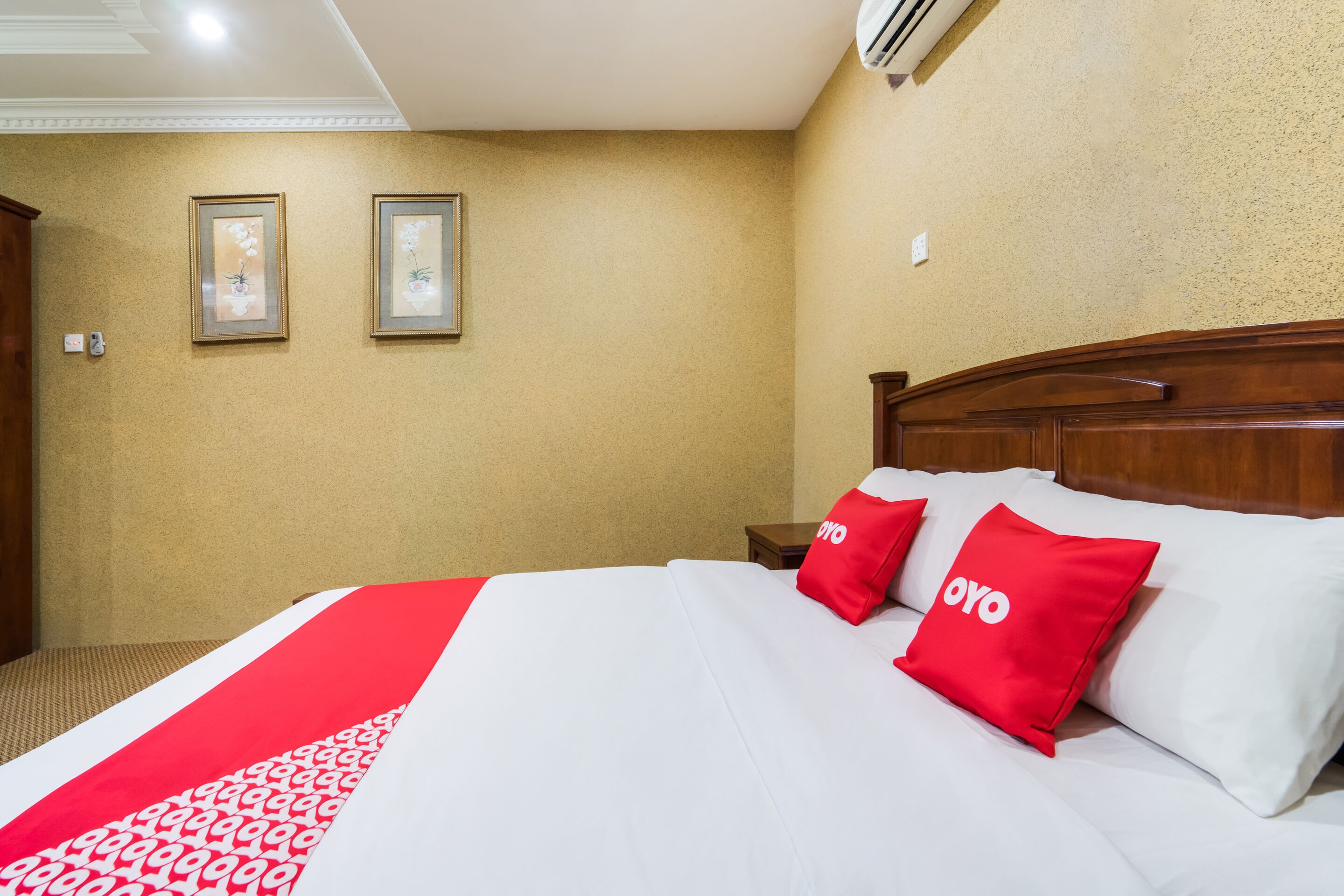 A hotel room with a large bed, two red pillows with the word "OYO" on them, a wooden headboard, and two framed pictures on the wall.