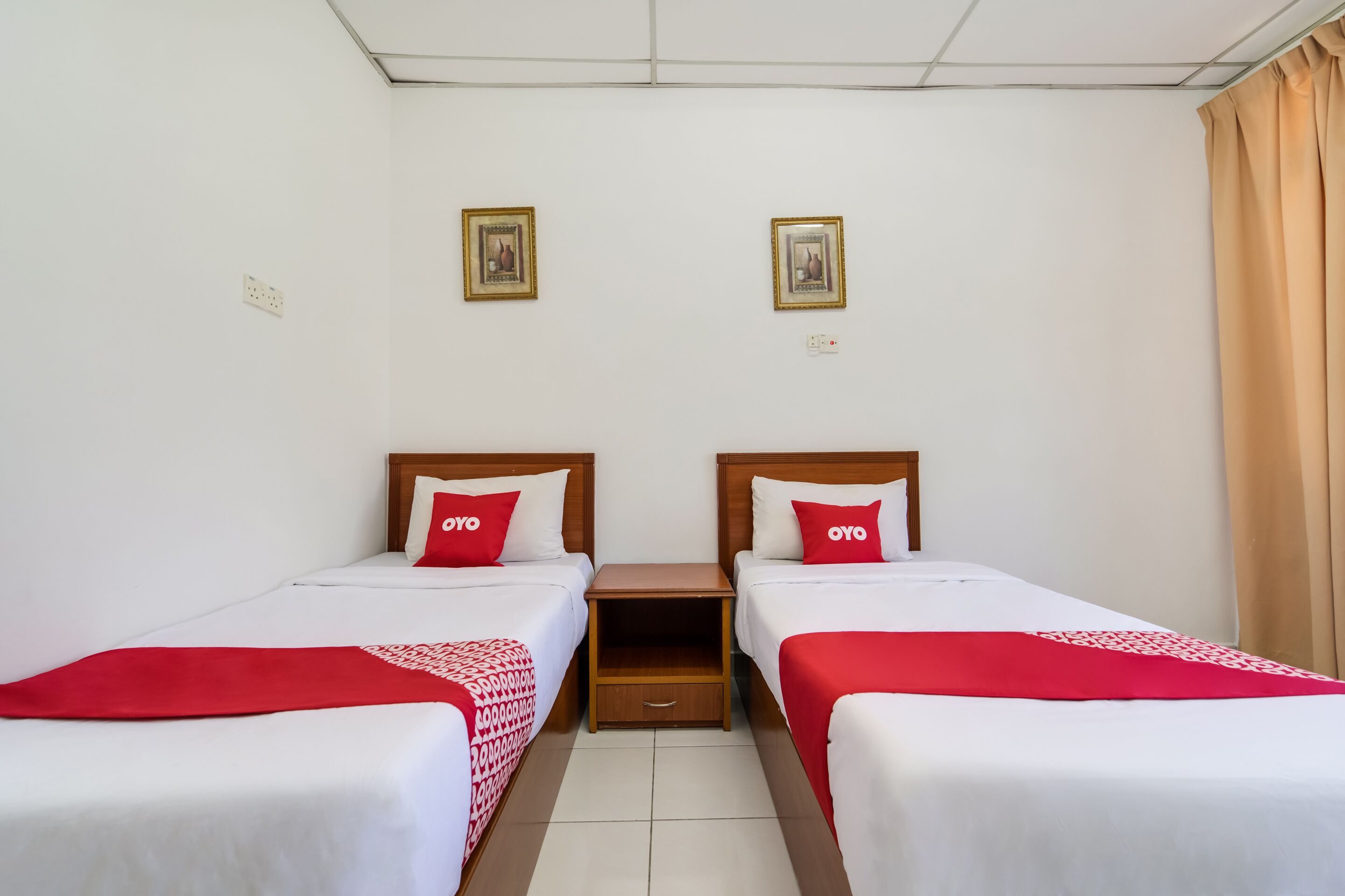 A hotel room with two single beds, each with a red and white bedspread and matching pillows. There are two framed pictures on the wall above the beds.