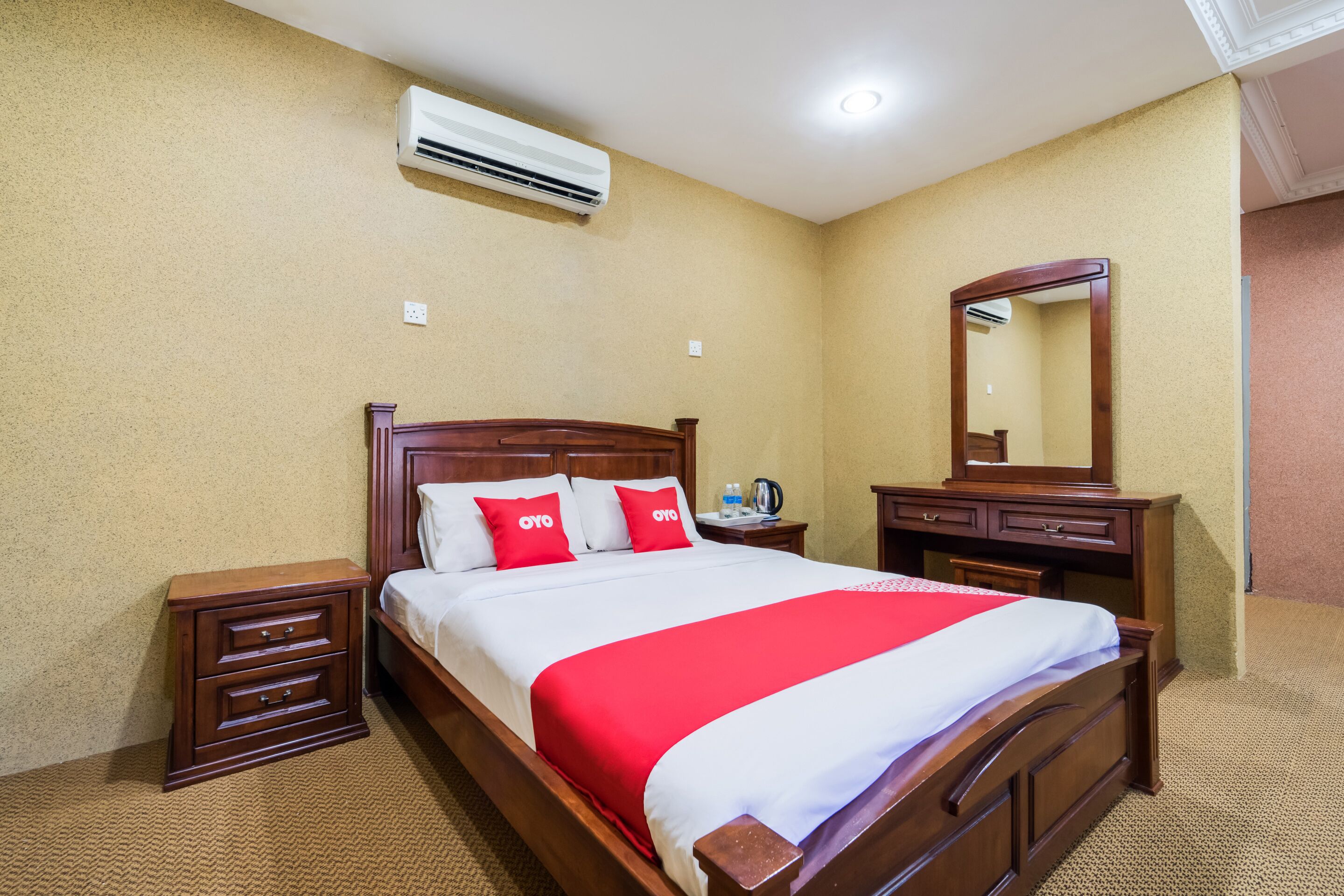 A hotel room with a wooden bed, a nightstand, a dresser, a mirror, and an air conditioning unit on the wall.