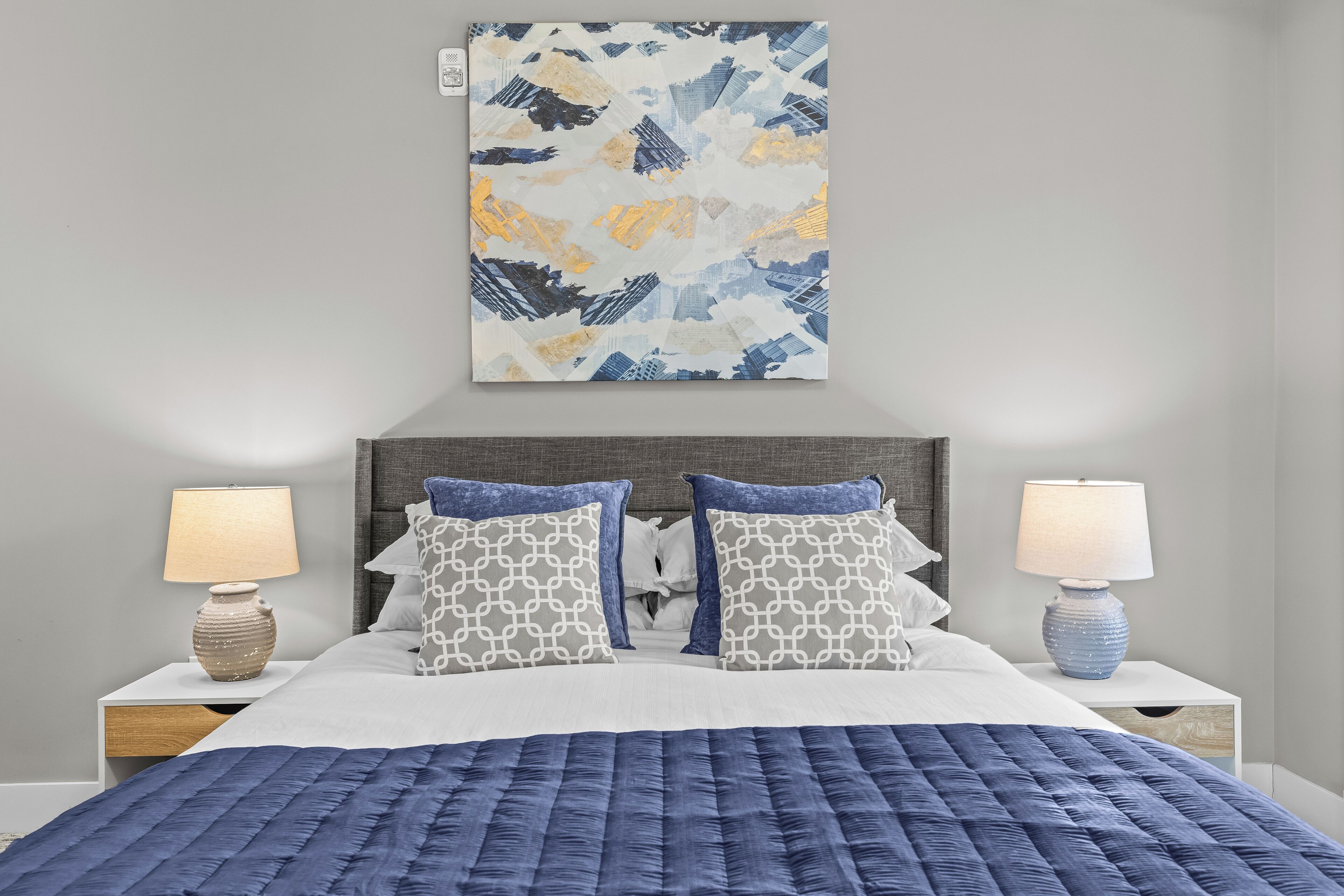 Una cama bien hecha con colcha azul, dos almohadas estampadas y un cuadro abstracto en la pared.