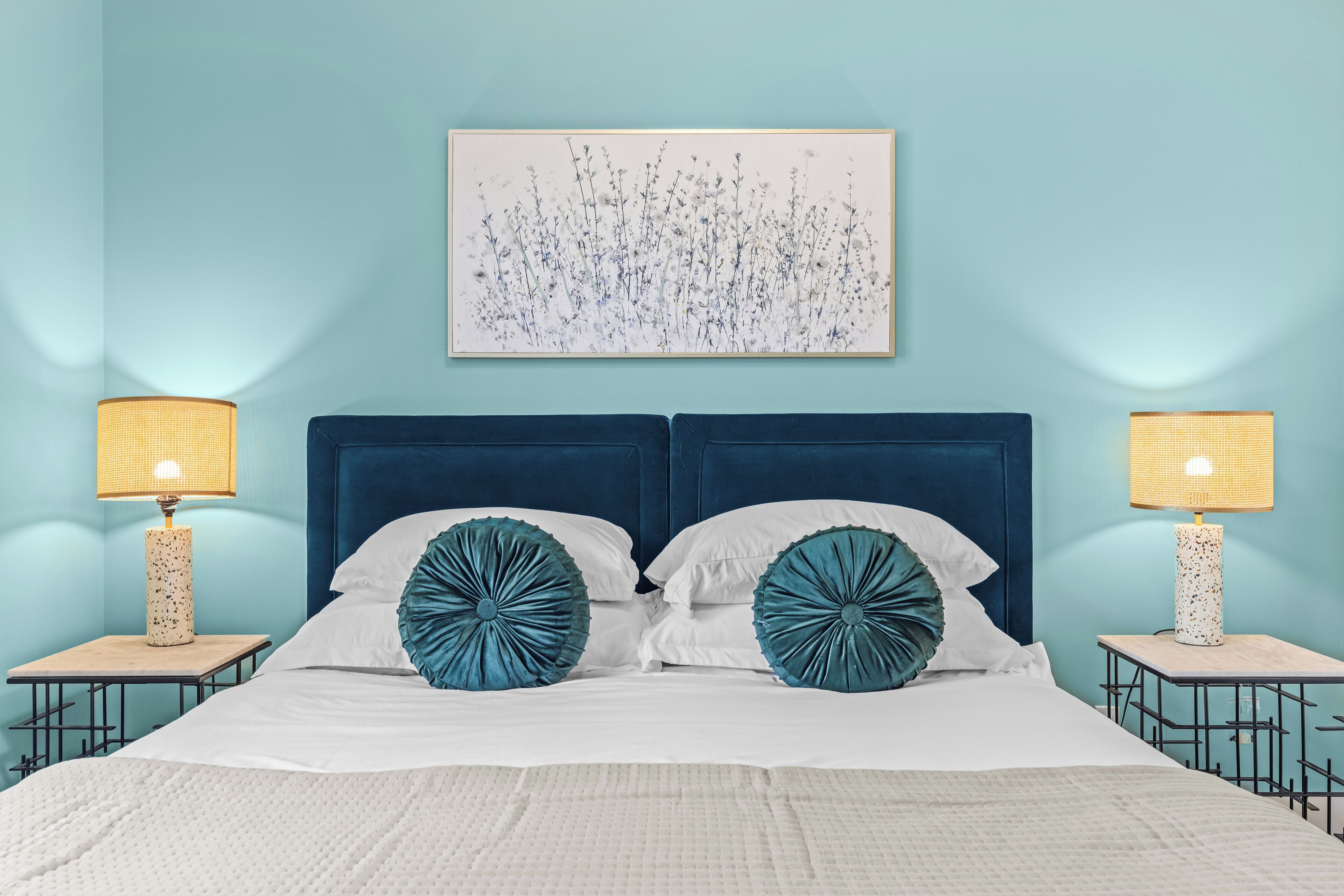 A bed with a blue headboard, white bedding, and teal decorative pillows. There are two bedside tables with lamps.