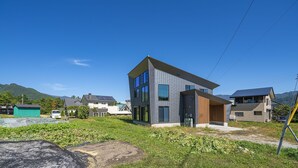 Una casa moderna de dos pisos con ventanas amplias y techo plano, rodeada de vegetación y colinas.