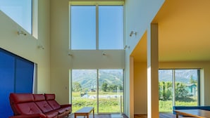 Amplia sala de estar con un sofá de cuero rojo, una mesa de centro de madera y una cama grande. La habitación cuenta con techos altos, ventanas amplias que ofrecen vistas de un paisaje montañoso y un diseño arquitectónico moderno.