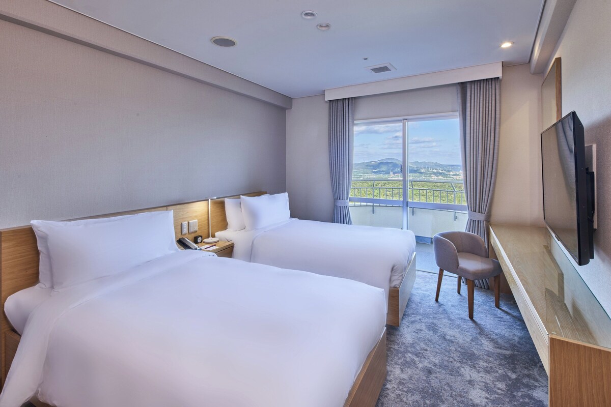 A hotel room with two beds, a desk, a chair, a TV, and a balcony with a city view.