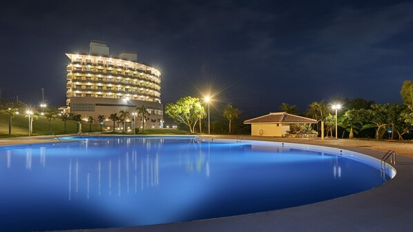 A swimming pool with a curved edge, surrounded by a well-lit area with palm trees and a building in the background. - ANA Crowne Plaza Resort Okinawa Uruma Hills by IHG (Uruma)