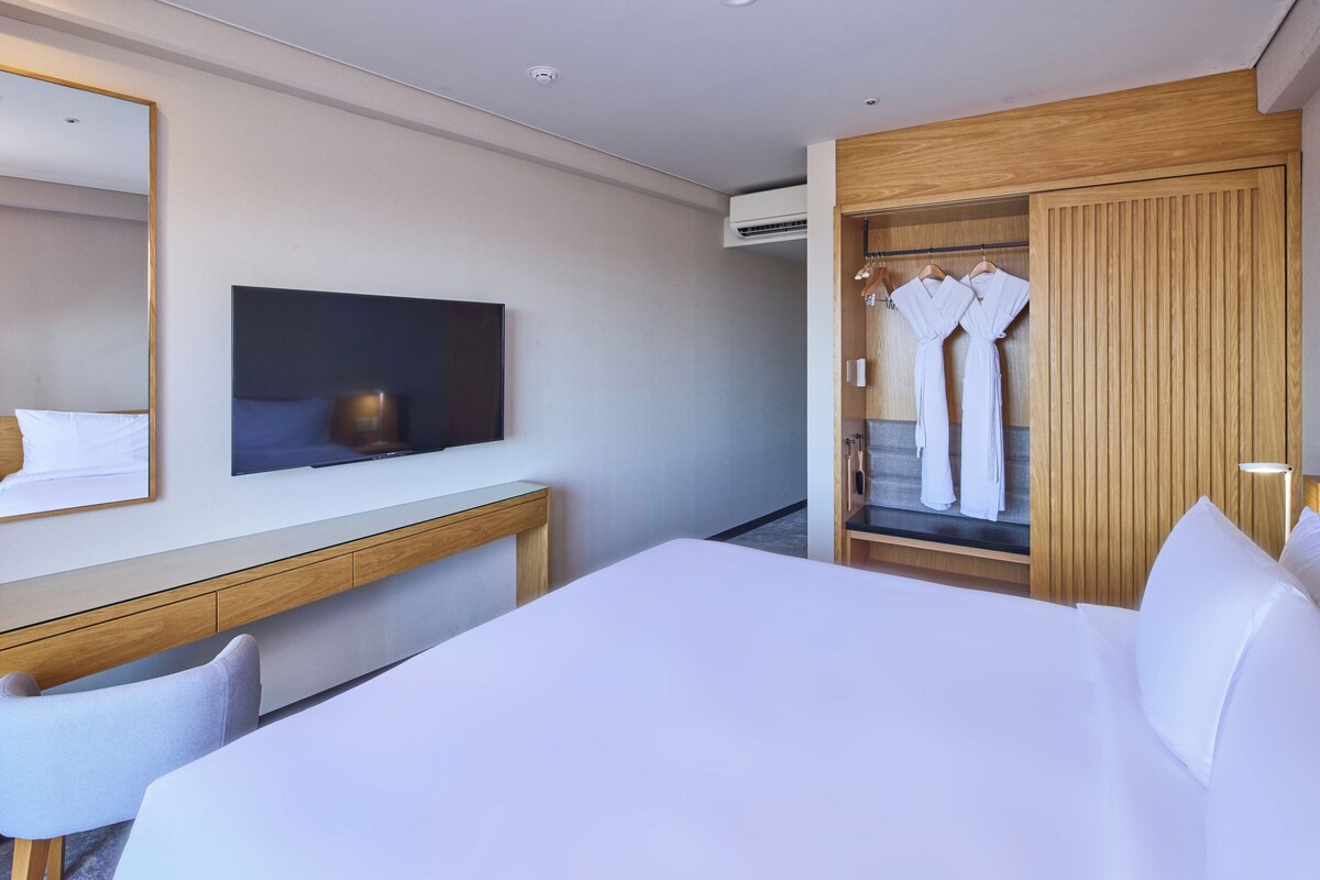 A modern hotel room with a large bed, a desk, a TV, and a wardrobe with robes.