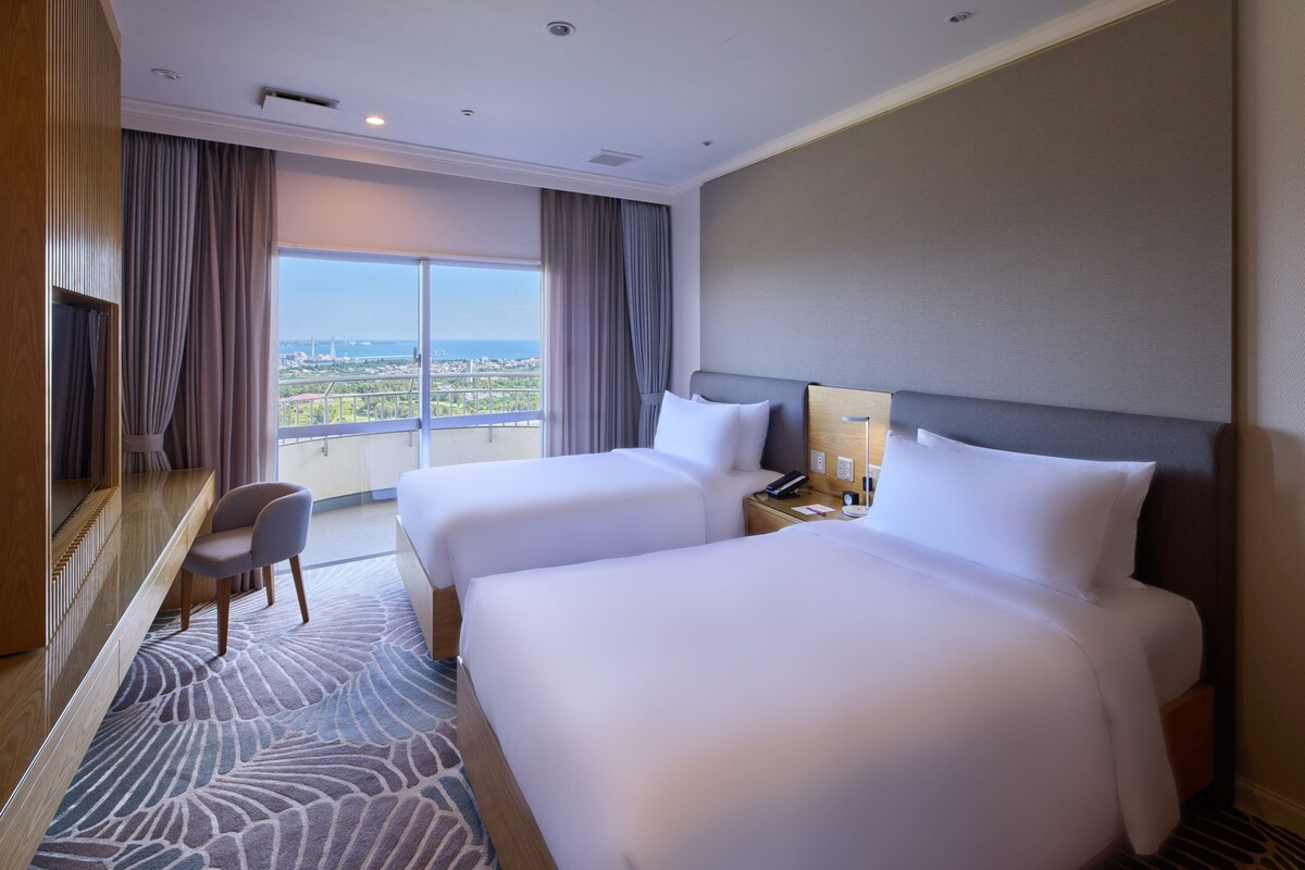 A hotel room with two beds, a desk, and a view of the city.