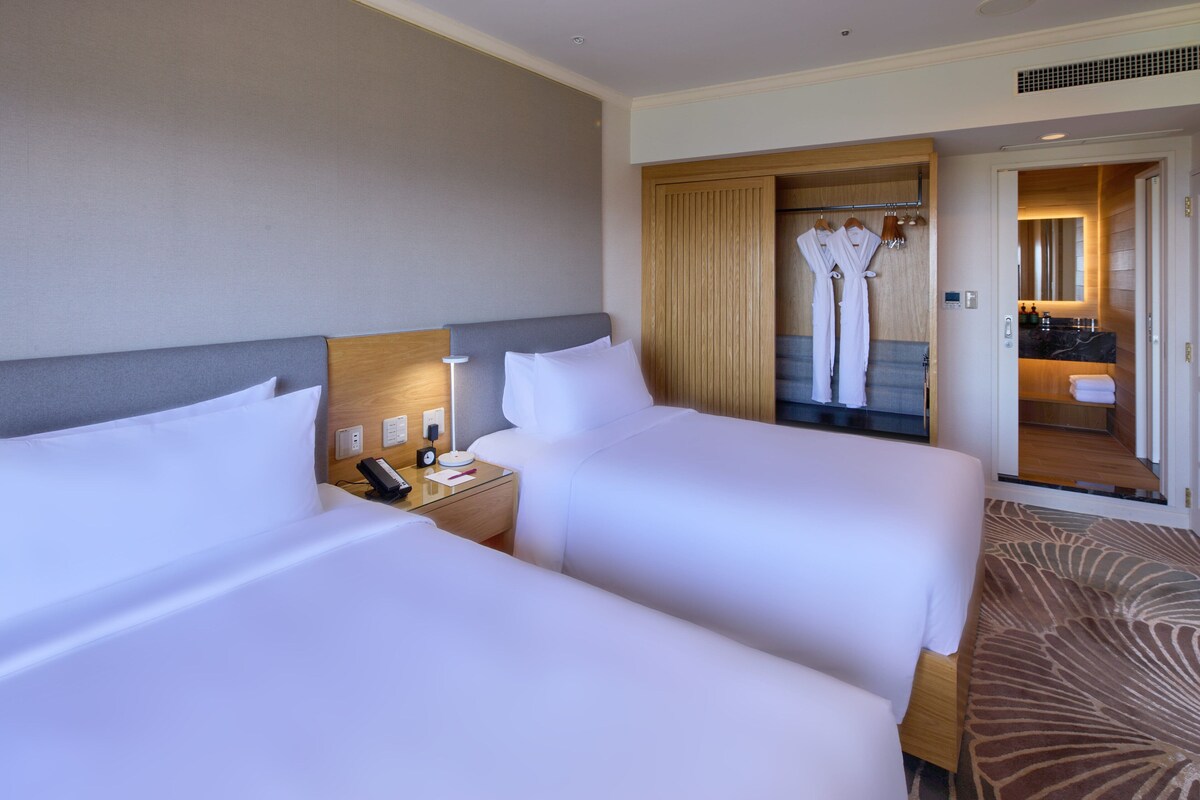 A hotel room with two beds, a bedside table with a lamp, a mirror, and a bathroom visible through an open door.