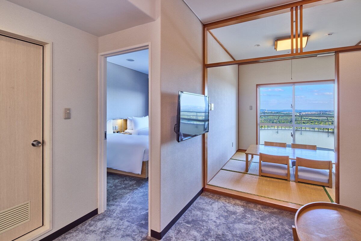 A hotel room with a bed, a TV mounted on the wall, a dining area with a table and chairs, and a large window offering a view of the outside.