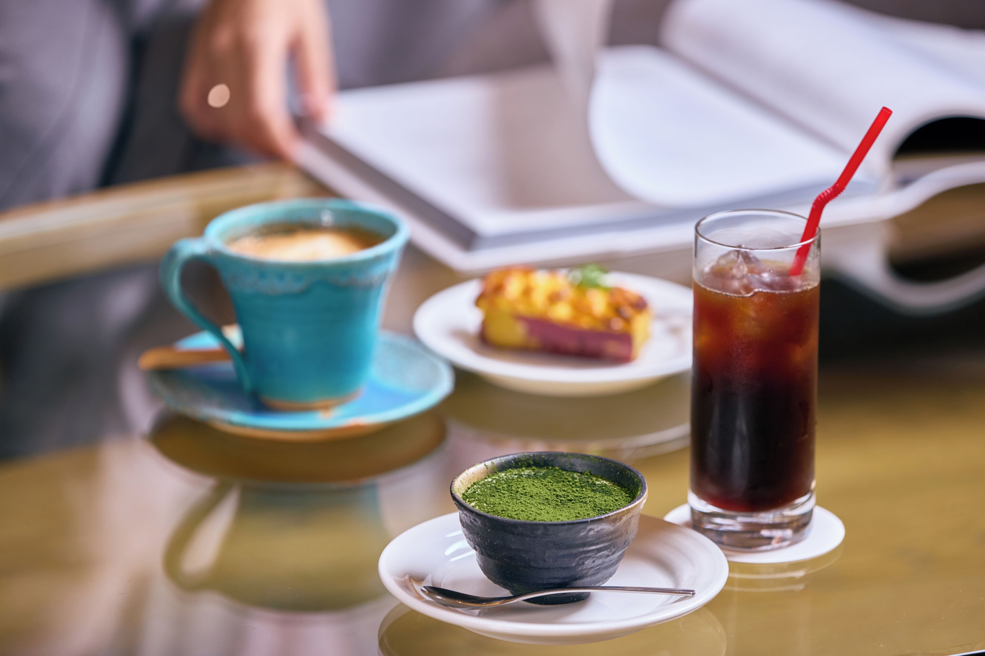 A table with a cup of coffee, a glass of iced tea, and a small bowl of green powder, possibly matcha, on a white plate.