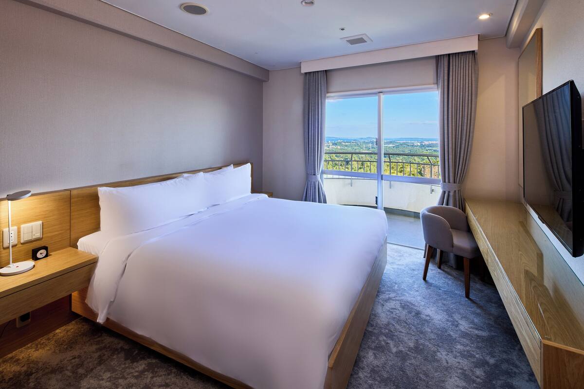 A hotel room with a large bed, a desk with a chair, a TV, and a balcony with a view.