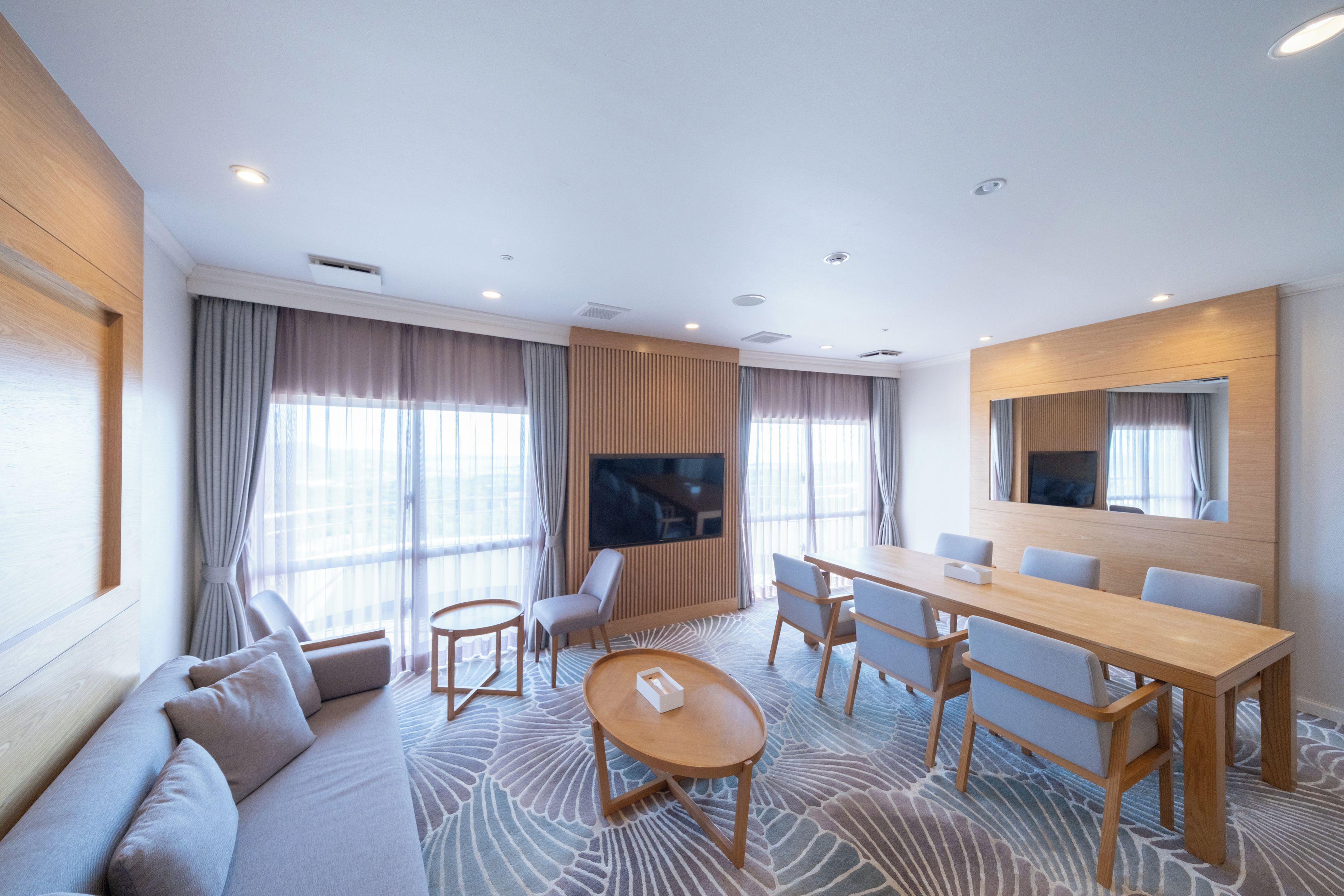 A hotel room with a sofa, a round table, a rectangular dining table, and a TV.