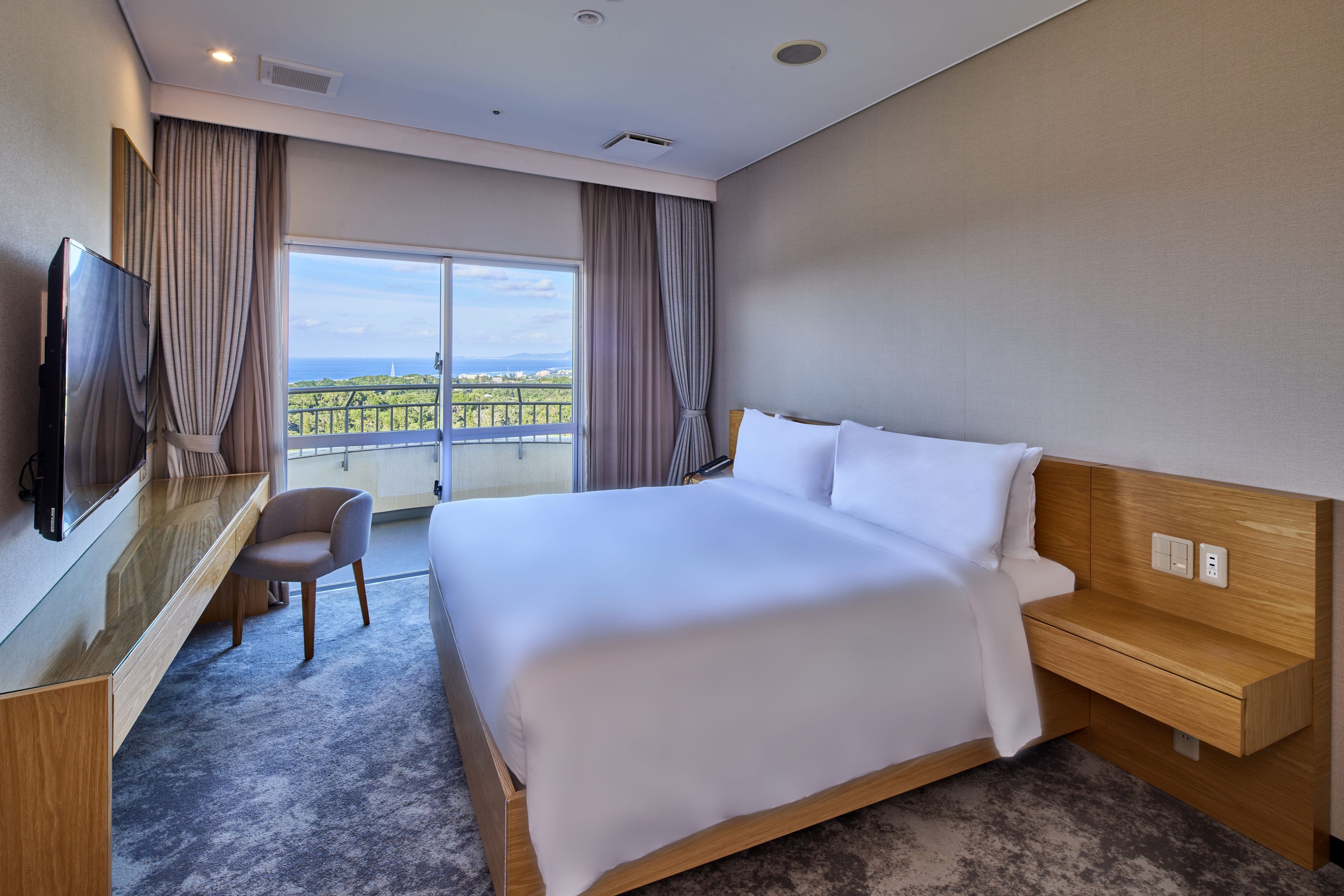 A hotel room with a large bed, a desk with a flat-screen TV, a chair, and a view of the cityscape through the window.